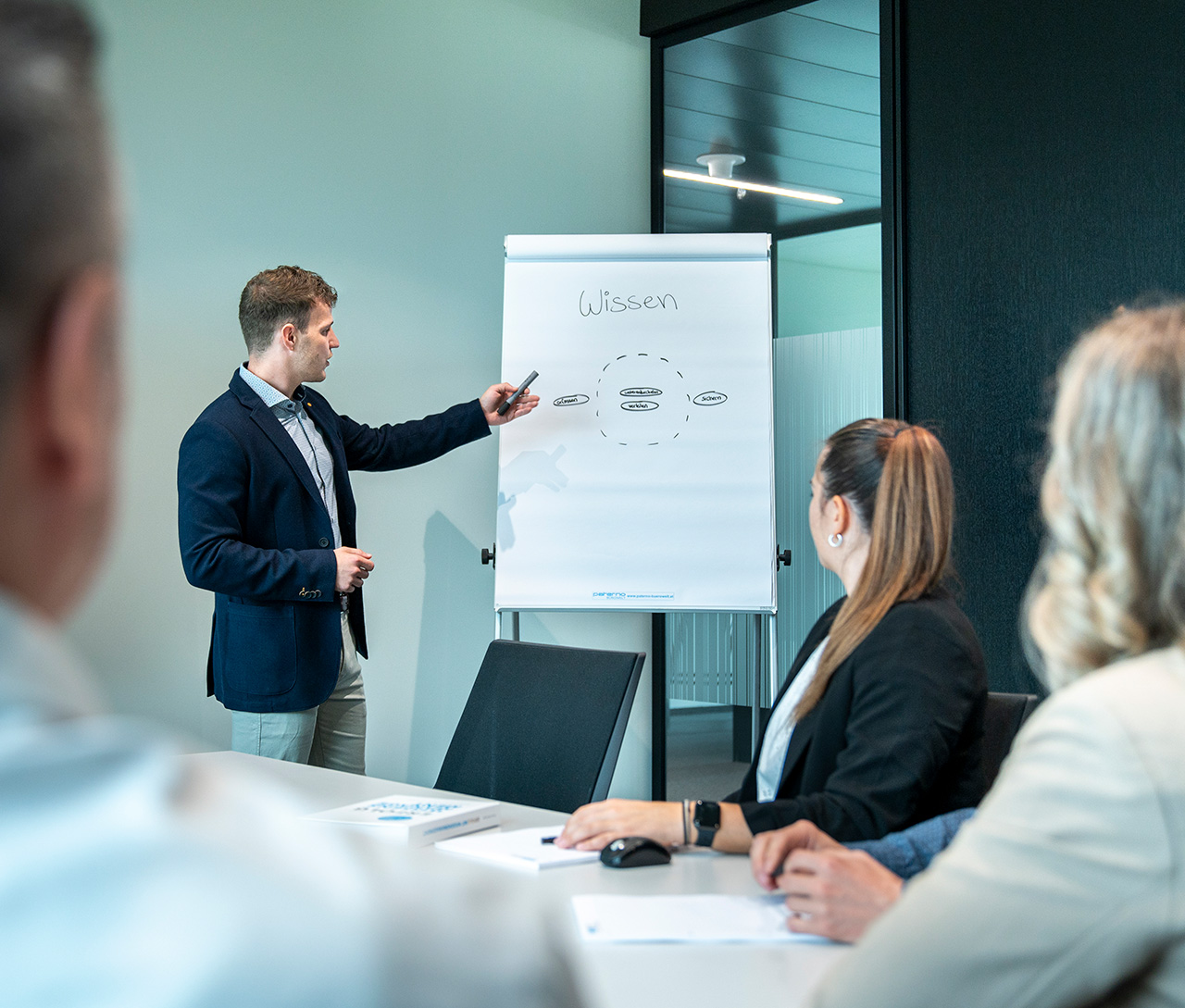 WBI-Academy_Zert-WM-Headerbild_1280x1080 Ein Mann zeigt drei Kollegen in einem Besprechungsraum ein Flipchart-Diagramm.