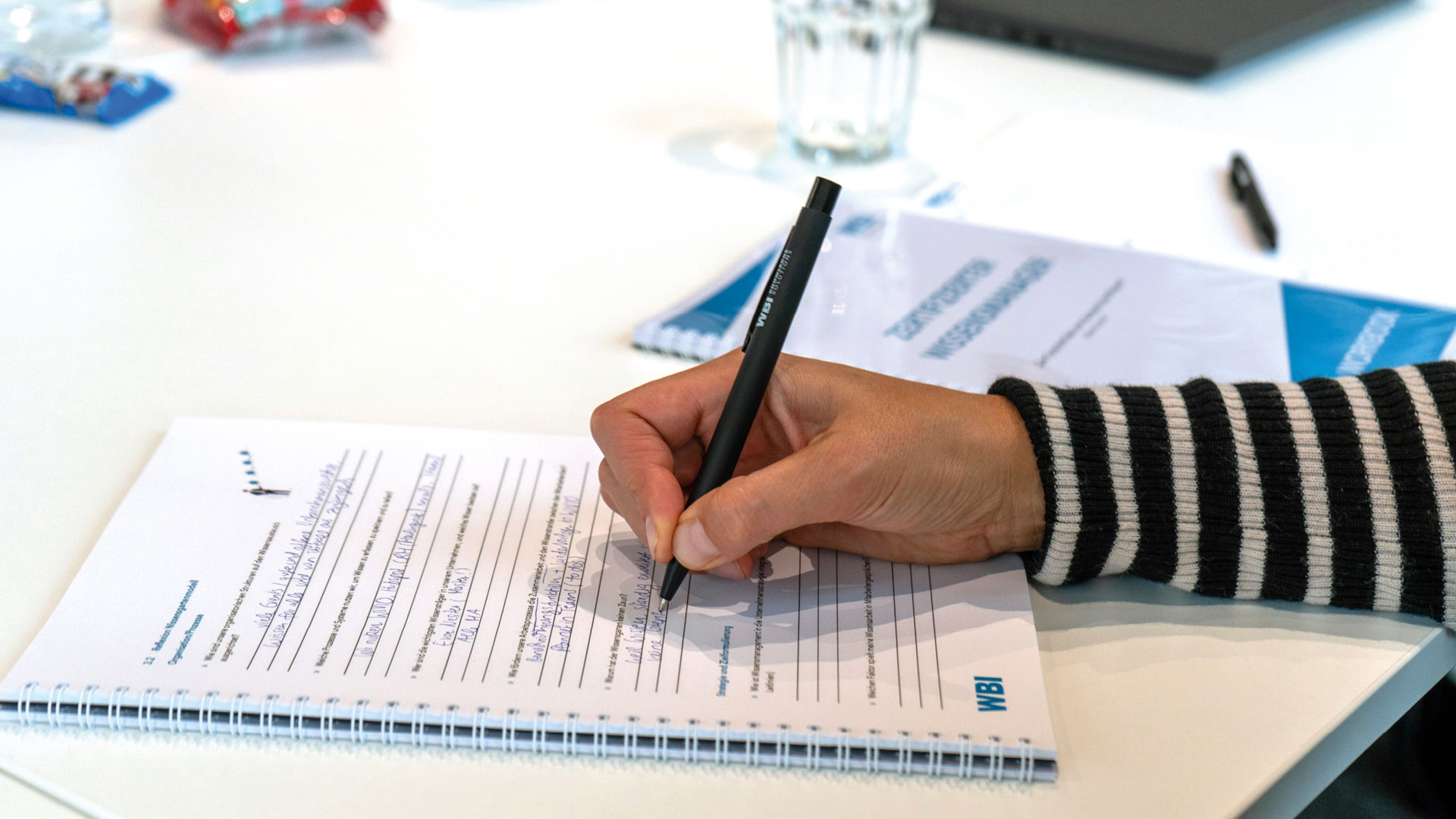 wbi-academy-zert-wm-mitschrift-web Eine Person schreibt an einem Schreibtisch in ein Notizbuch.