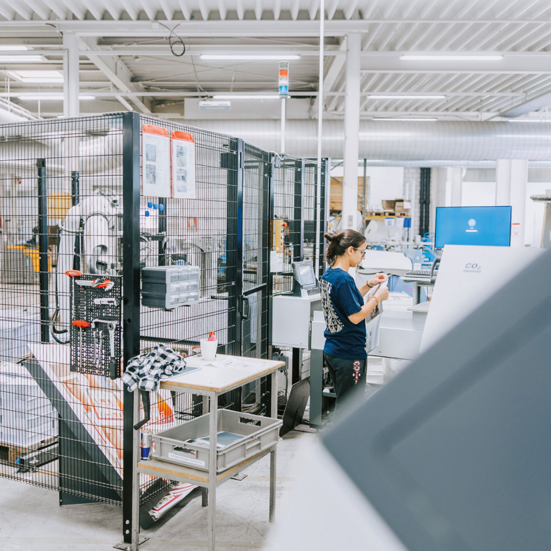 wbi-casestudy-VVA-produktion-web Das Bild zeigt die Produktion der VVA in Dornbirn. Eine Mitarbeiterin steht an einer Maschine und kontrolliert gerade eine Drucksorte, die in der Vorarlberger Verlagsanstalt produziert wurde. Im Hintergrund ist ein Roboterarm sichtbar, der die Mitarbeiter in der Druckerei unterstützt.