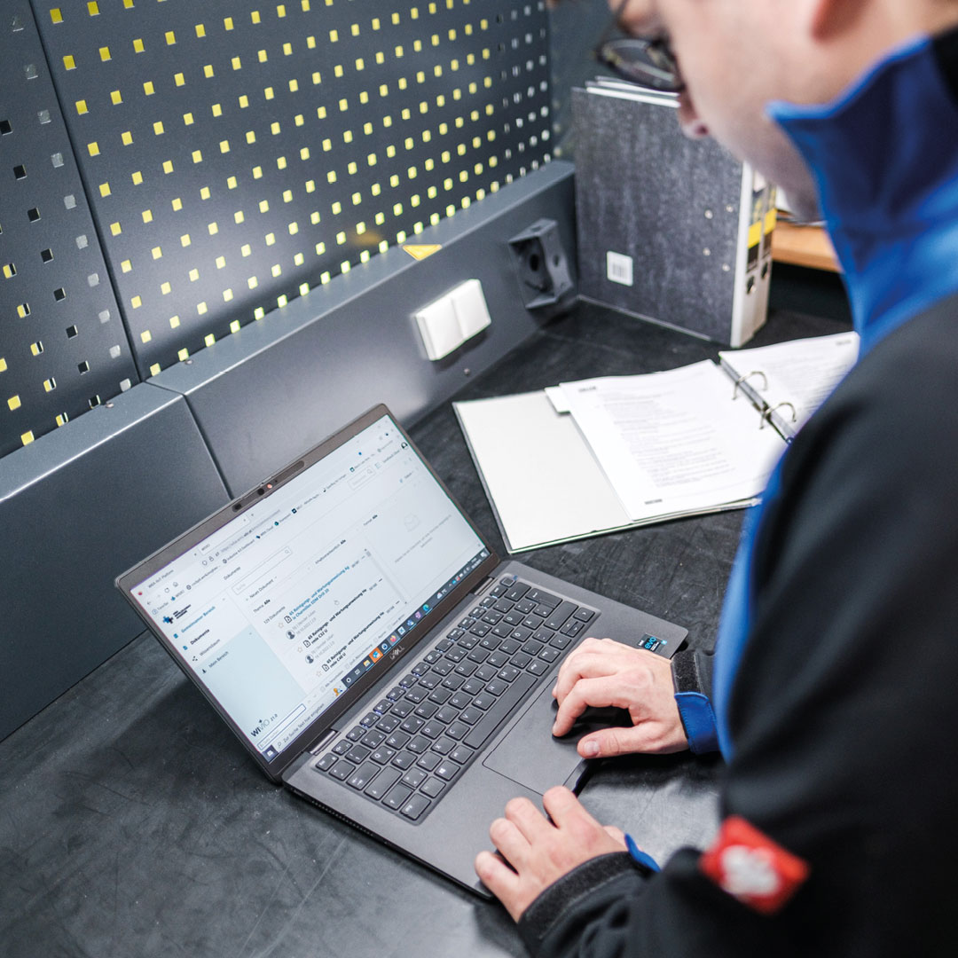 wbi-casestudy-wba-laptop-web Ein Mitarbeiter der WBA Aachender Werkzeugbau Akademie steht an einem Schreibtisch mit einem Laptop, mit Dokumenten und einem Ordner daneben.