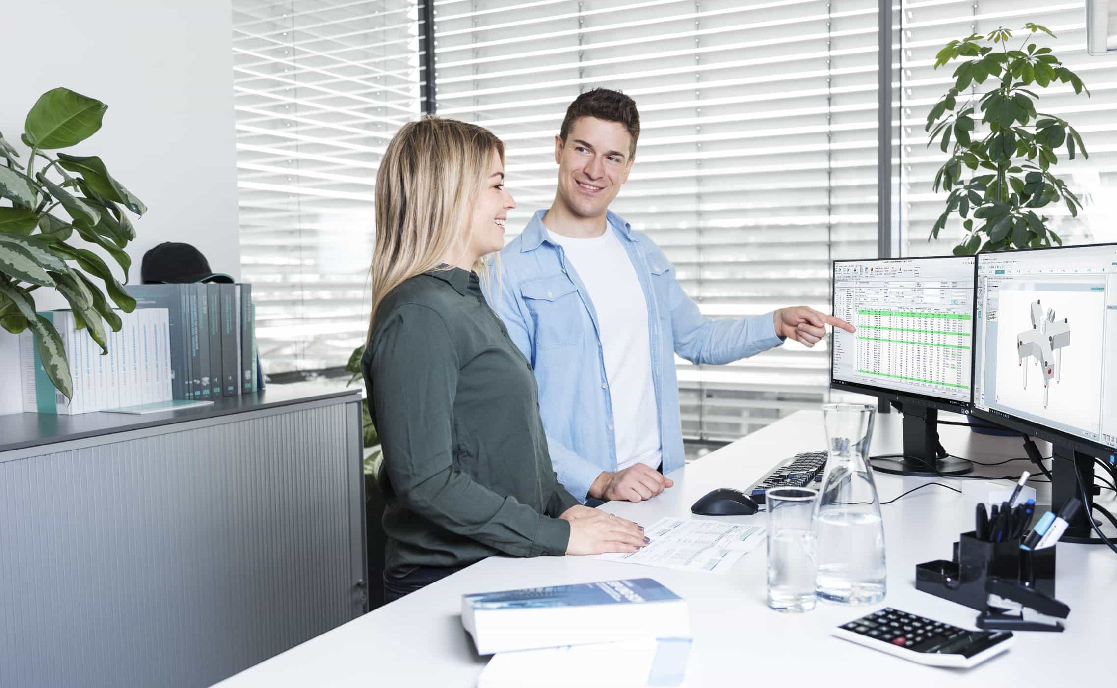 Zwei Kollegen lächeln und besprechen Daten auf Computerbildschirmen im Büro.