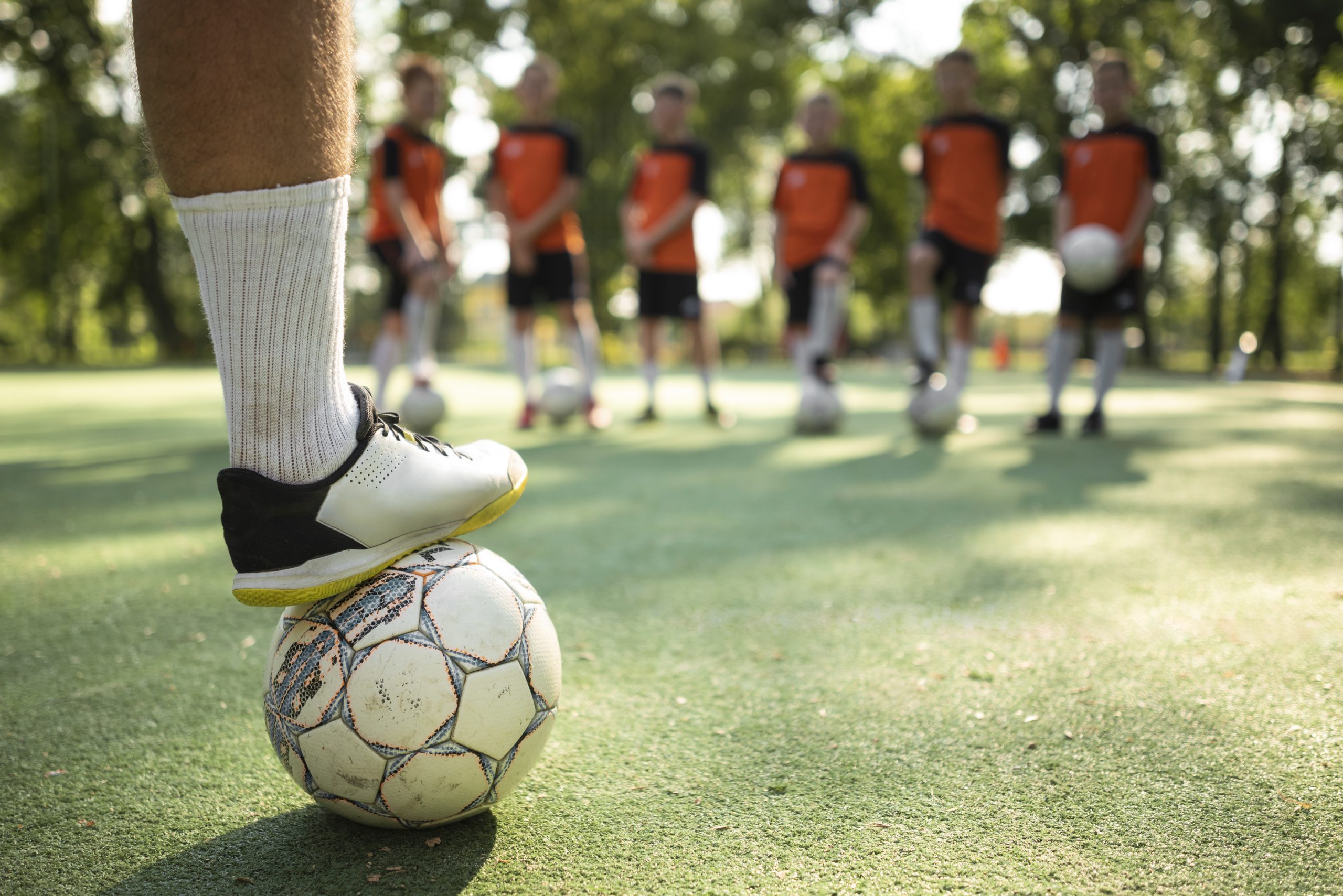 wissensmanagement-fur-vereine-verbande-und-service-clubs Das Bild zeigt im Vordergrund ein Bein mit weißen Socken und Turnschuhen auf einem Fußball ruhend. Im Hintergrund verschwommen 6 Kinder im Fußballtrikot mit einem Ball in der Hand oder am Boden.